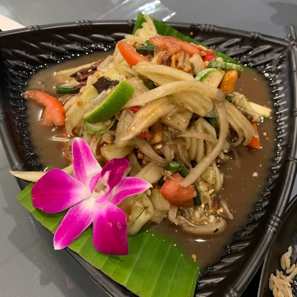 Thai papaya salad with tomatoes, lime, and a purple orchid garnish on a black plate.