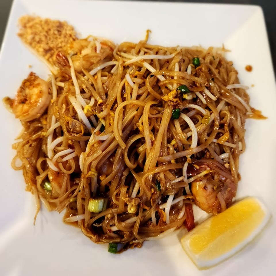 A white plate of Pad Thai noodles with shrimp, bean sprouts, crushed peanuts, and a lemon wedge.