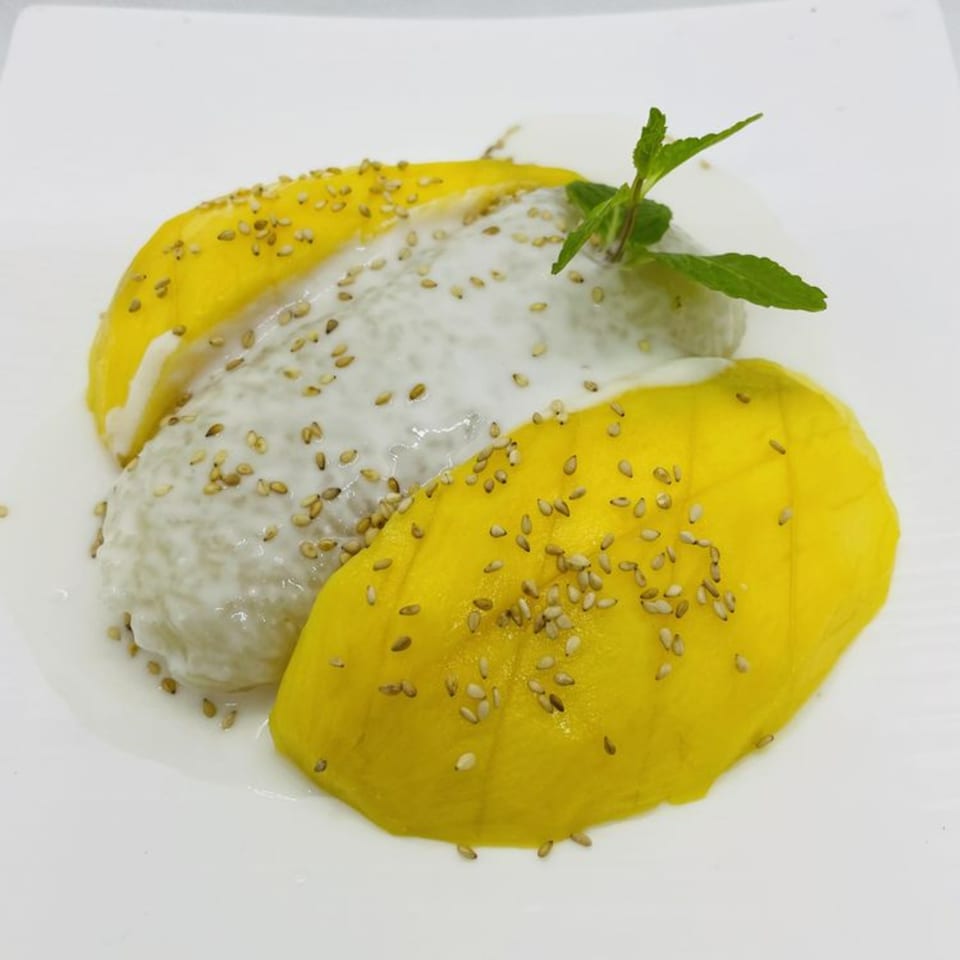 Thai mango sticky rice dessert with coconut milk, sesame seeds, and mint on a white plate.